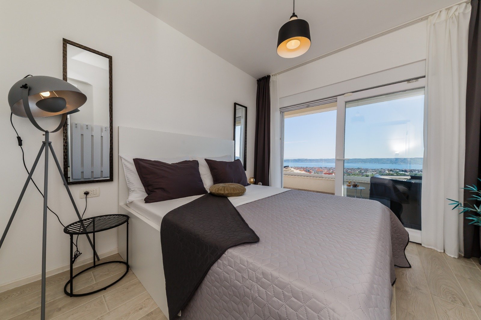 Bedroom with grey bedding, a tripod floor lamp and glass balcony doors overlooking the sea.