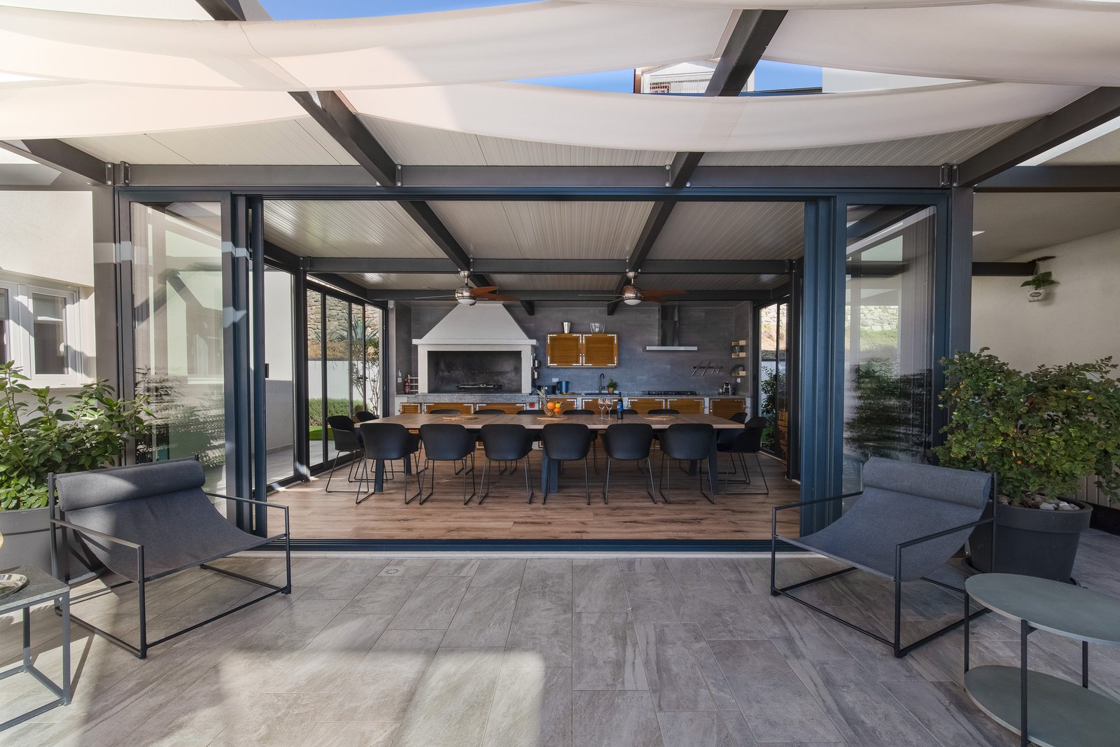 Open glass doors connect shaded courtyard chairs with the covered dining pavilion, long table and barbecue.