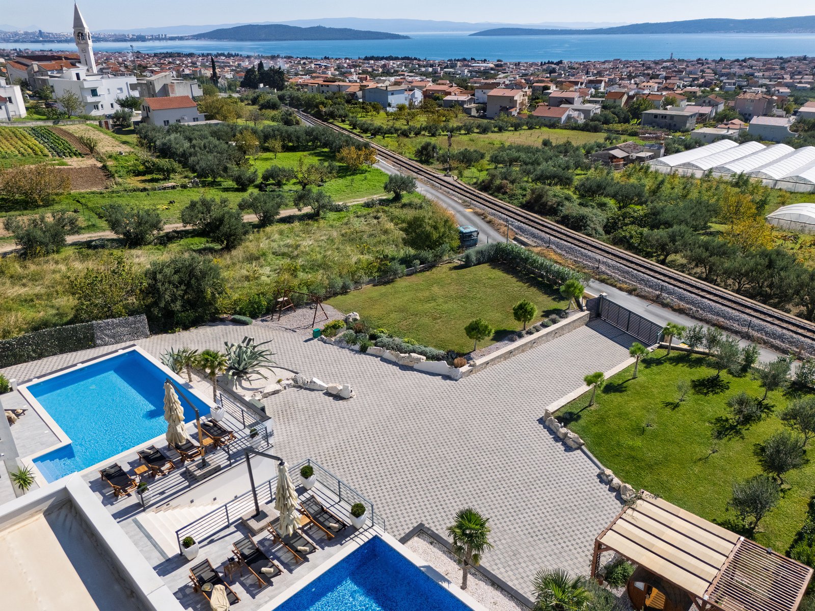 Elevated view over the estate's pool terraces, lawns, swing frame and courtyard toward nearby railway tracks, coastal rooftops and the sea.
