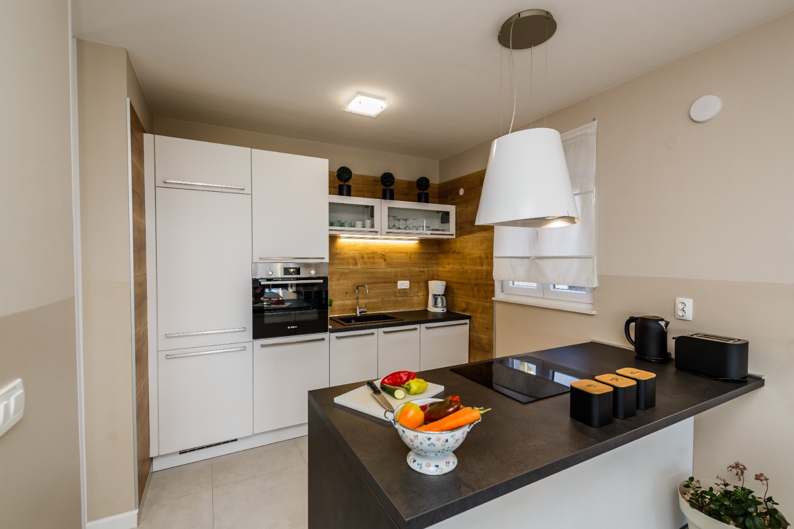 White kitchen with dark worktops, a wood-effect backsplash, built-in oven and hob on the peninsula.