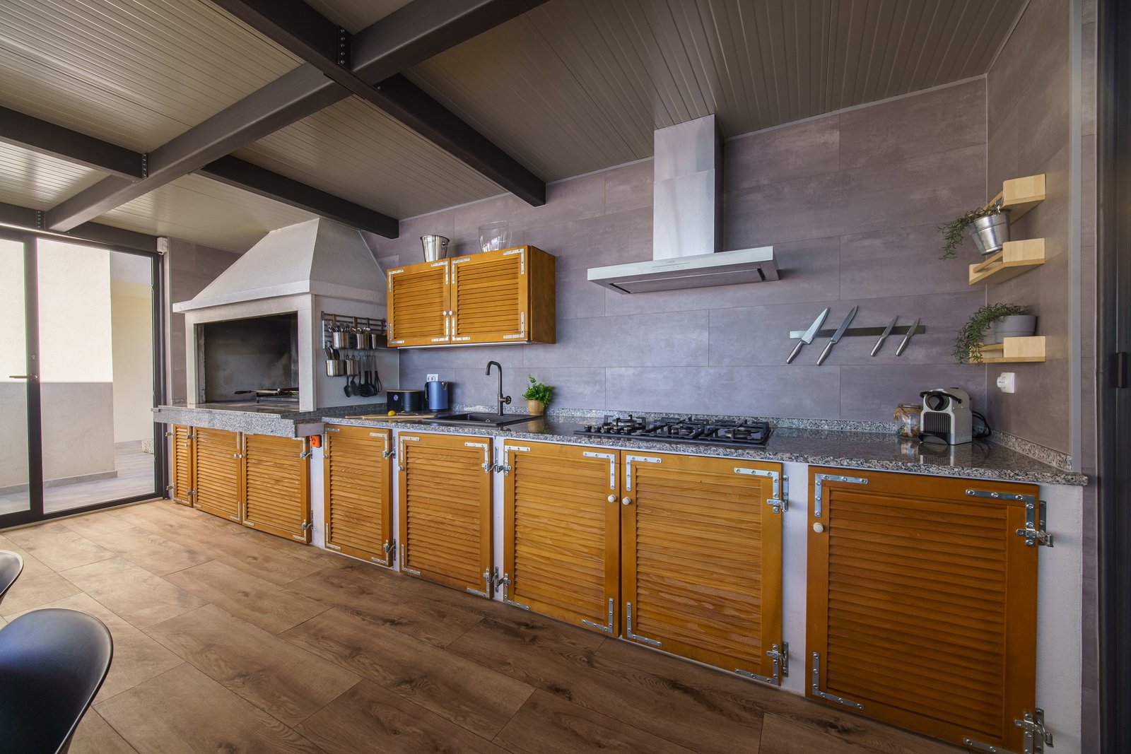 Kitchen in the covered pavilion with a built-in barbecue, gas hob, sink, wooden cabinets and coffee machine.