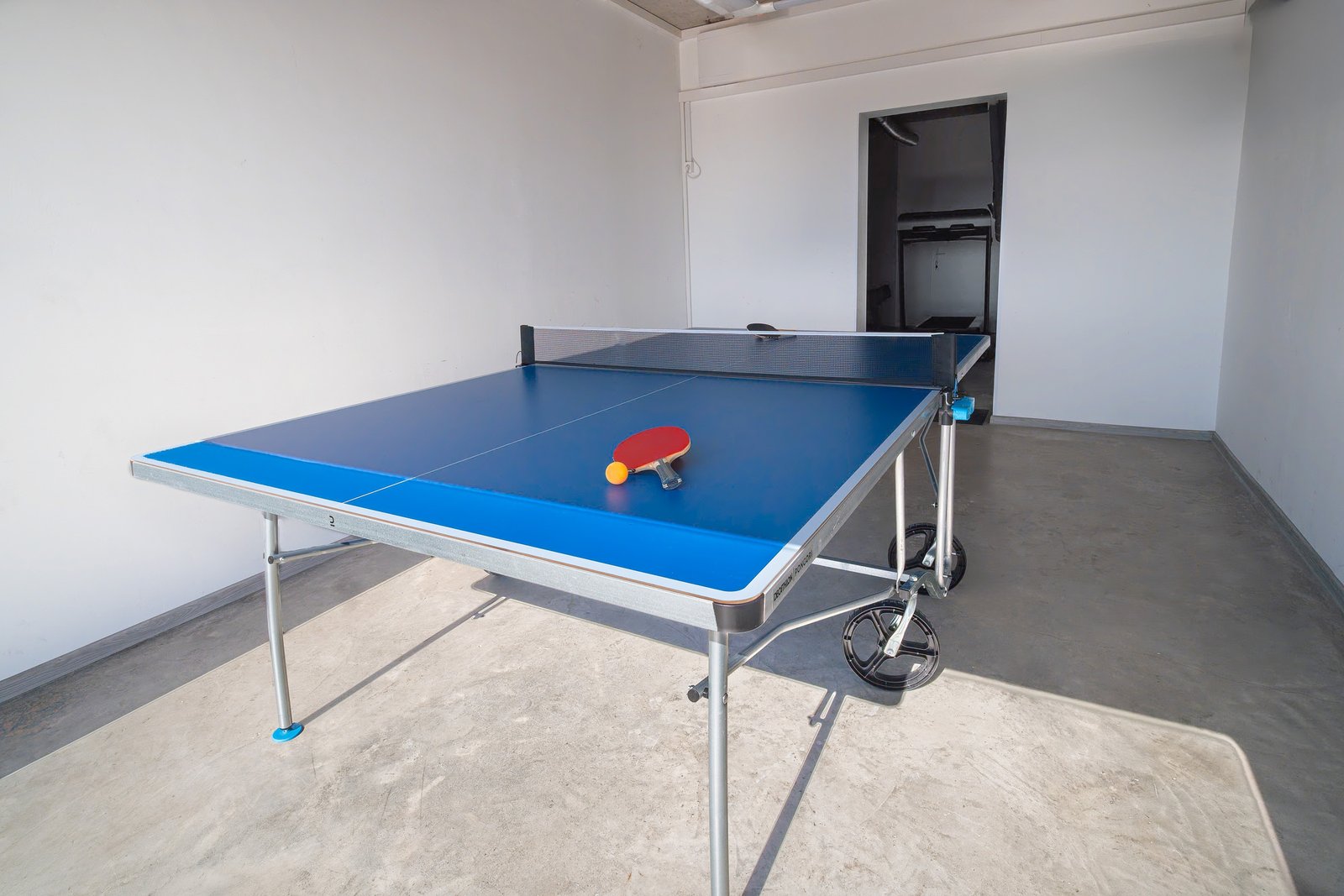 Blue indoor table tennis table with a net, paddles and an orange ball.